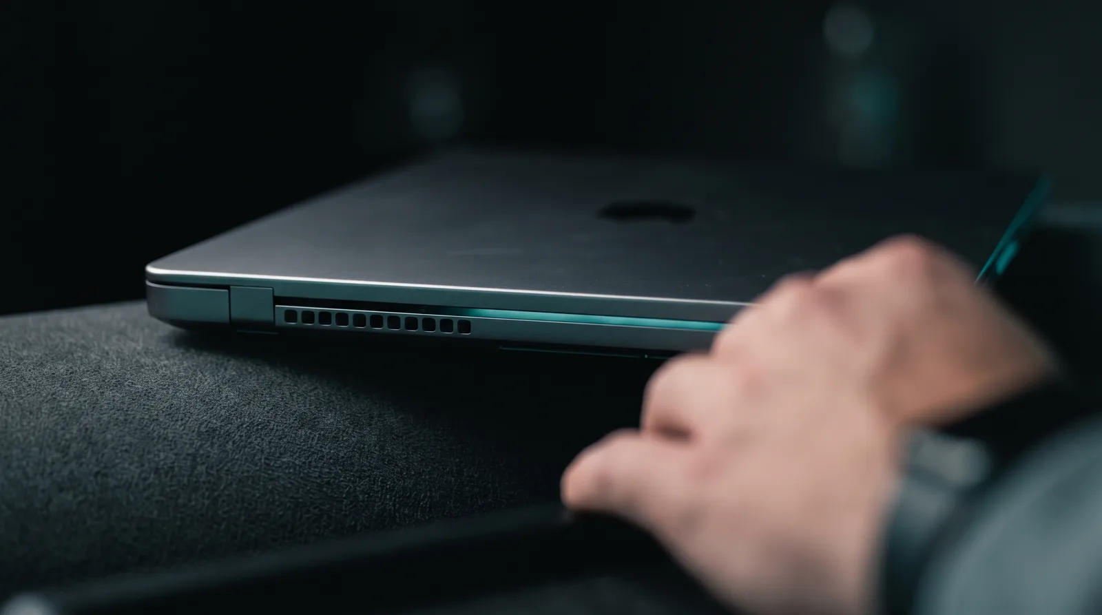 Close-up of an open Mac laptop's aluminium edge with cyan-teal light seeping from the display gap on a dark background.