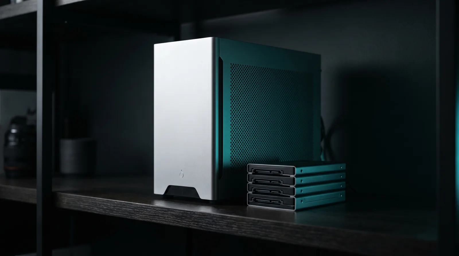 A silver compact desktop workstation and a stack of solid-state drives lit by cyan-teal accent light on a dark shelf.