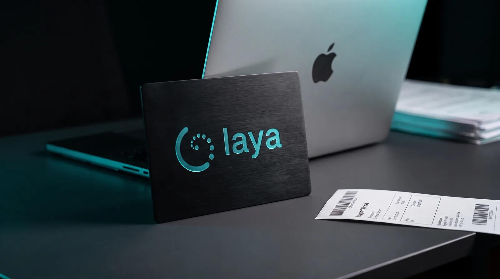 Dark editorial tech scene of an Apple laptop and a metal plate bearing the Laya decision-model logo beside a printed support ticket