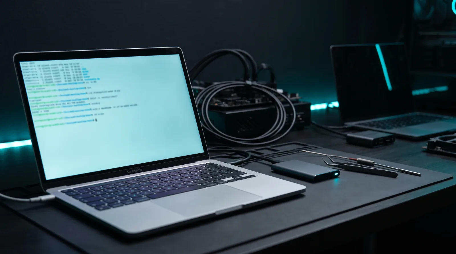 Silver laptop on a dark workbench with an external SSD and a thermal probe beside it, lit by cyan terminal glow, illustrating a local LLM drafting benchmark on Apple Silicon.
